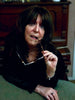 A woman with long dark hair leans on a green surface, holding eyeglasses near her mouth and gazing thoughtfully. She wears jewelry inspired by crystal magick and has The Witch's Oracle, 2nd Edition by Schiffer Publishing, Ltd. beside her.