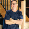 A man with short brown hair and glasses, wearing a navy blue t-shirt and green pants, stands in a workshop by wooden stairs, ready to share his Pinball expertise from Schiffer Publishing, Ltd.