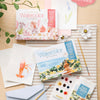 A flat lay of Schiffer Publishing, Ltd.’s Watercolor the Easy Way Paint-Your-Own Watercolor Cards Coastal kit, art supplies, floral and lobster illustrations, paint palette, brushes, a flower, envelope, notecards, and a plant on a wooden table.