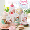 A bright, colorful display features the Schiffer Publishing, Ltd. Crochet Cafe Amigurumi Kit Cake Slice among crocheted desserts and yarn, set on pastel plates with a floral teapot and a "Collect the whole set!" sign in the background.
