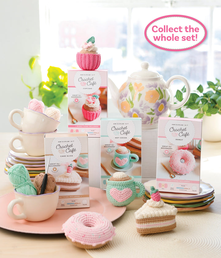 A bright, colorful display features the Schiffer Publishing, Ltd. Crochet Cafe Amigurumi Kit Cake Slice among crocheted desserts and yarn, set on pastel plates with a floral teapot and a "Collect the whole set!" sign in the background.