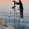 A man on a step ladder in shallow ocean water adjusts a camera at sunset, with rocks nearby and the calm sea under a pink sky—capturing scenes for Midwest Home by Schiffer Publishing, Ltd.