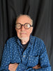 An older man with a gray beard and round glasses stands with arms crossed in a blue polka-dot shirt before a black curtain, embodying the spirit of "The Story of the Blues in 50 Songs" by Schiffer Publishing, Ltd.