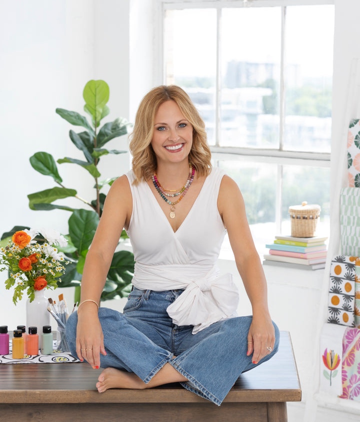 A smiling blonde woman sits cross-legged on a wooden table in a bright room with art supplies, flowers, and plants. She wears a white top, blue jeans, colorful necklaces—and has the Schiffer Publishing Watercolor the Easy Way Paint-Your-Own Watercolor Cards Garden kit nearby.