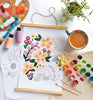 The ColorMaps Paint-by-Number Beautiful Blooms by Schiffer Publishing, Ltd. is in progress on white paper, surrounded by paintbrushes, watercolor paints, bottles, a palette, coffee cup, plant, and another floral sketch.