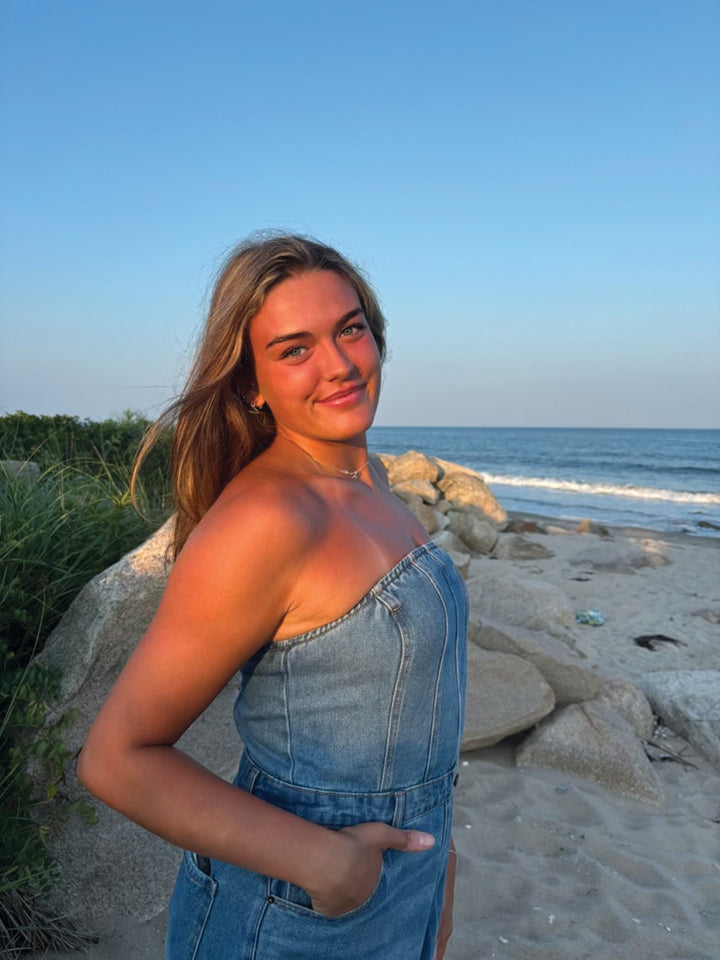 A young woman stands on a sandy beach at sunset, smiling warmly with her hands in her pockets, surrounded by nature patterns inspired by the Wild & Whimsical Woodland Coloring Book from Schiffer Publishing, Ltd.