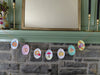 The "Pumpkin Patch Pals" garland by Schiffer Publishing, Ltd. features charming illustrations of pumpkins, mushrooms, leaves, and donuts strung across a stone fireplace mantle for an autumn-inspired display.