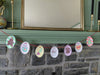 A string of oval-shaped paper ornaments from the "Christmas Market Crew" by Schiffer Publishing, Ltd., inspired by paper craft coloring book designs and adorned with festive holiday art, hangs on a red string across a gray stone fireplace beneath a green mantel and large mirror.