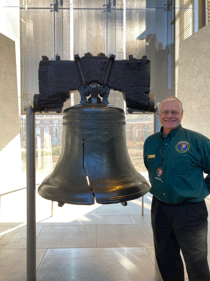 A smiling man in a green shirt stands beside the Liberty Bell, its famous crack visible, inside Independence National Historical Park. Sunlight streams through windows behind them. Product by Schiffer Publishing, Ltd.