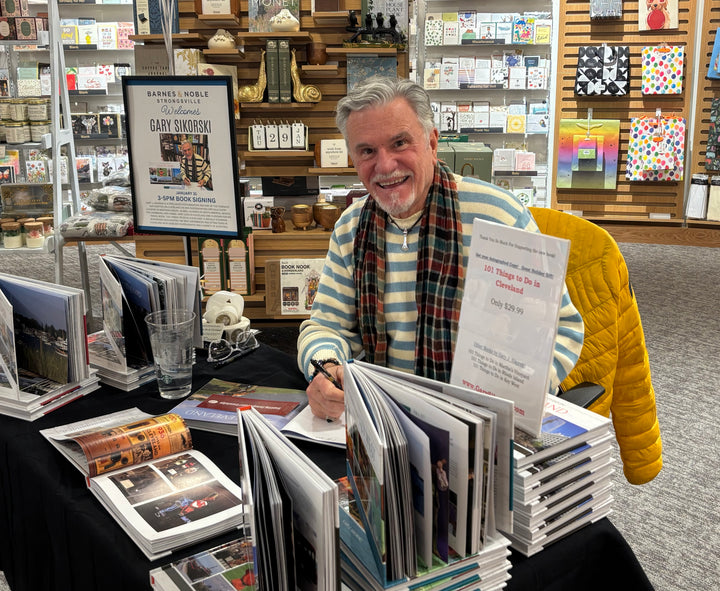 A smiling man in a striped sweater and plaid scarf signs copies of "Detroit" by Schiffer Publishing, Ltd. at a table surrounded by colorful Motor City souvenirs, promotional signs, and shelves filled with gifts and cards in a bookstore.