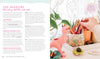 A desk with art supplies—colored pencils in a cup, the "Good Vibes Art Club Learn to Draw" book by Schiffer Publishing, Ltd., sketchbooks, a pink dinosaur figurine, small décor items, and a leafy plant—beside a window with lace curtains and a round mirror.