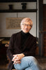 A man with light hair and glasses smiles in a traditional Japanese-style room with wooden walls made using joinery, next to "Traditional Japanese Woodworking" by Schiffer Publishing, Ltd., wearing a black turtleneck, brown jacket, and jeans.