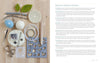 A flat lay of Schiffer Publishing, Ltd.'s "Needle Felting Through the Seasons" book and essential tools—felted wool balls, felting needles, safety eyes, yarn and circular needles, wool blocks, stitch markers, and templates—on a wood surface.