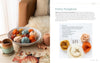 A bowl of handmade wool pumpkins in autumn hues sits near yarn, knitting needles, and a mug. Beside it is "Needle Felting Through the Seasons" by Schiffer Publishing, Ltd., featuring step-by-step cozy felting instructions.
