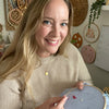 A smiling woman with long blonde hair holds the Sunny Stitches Embroidery Kit Sunshine Pines by Schiffer Publishing, Ltd., working on colorful stitches in a cozy craft room decorated with nature embroidery art on the wall behind her.