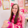 A woman with long brown hair, wearing a pink and red striped sweater, sits smiling in a colorful, craft-themed room adorned with art, paper flowers, cheerful signs, and whimsical stickers from Schiffer Publishing’s A Day in Cozy Town: The Bookshop Coloring and Sticker Book.