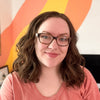 A woman with wavy brown hair and glasses smiles at the camera, wearing a peach shirt. Behind her, bold orange and yellow stripes brighten the white wall, capturing the playful spirit of a child’s painting as seen in the Schiffer Publishing, Ltd. I Heart Art Forest Friends Watercolor Paint Pad.