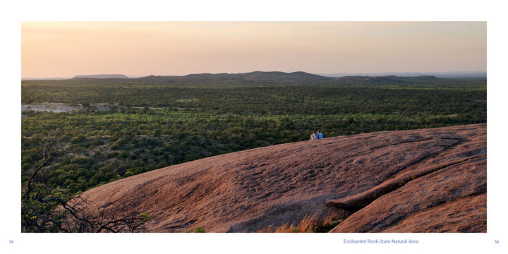 Two people sit on a large, rocky outcrop overlooking the Texas Hill Country at sunset.