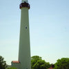 A tall, white lighthouse with a red top stands among trees and small buildings under a blue sky—a highlight featured in Cape May Walking Tours by Schiffer Publishing, Ltd.