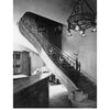Black and white photo of an elegant spiral staircase with Art Deco ironwork inspired by French metalsmith Edgar Brandt, a large chandelier above, bookshelves on the left, and framed art above the stairs. Product: "Edgar Brandt" by Schiffer Publishing, Ltd.