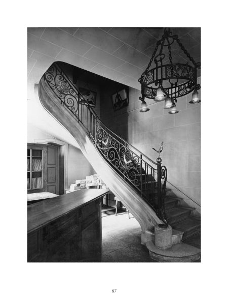 Black and white photo of an elegant spiral staircase with Art Deco ironwork inspired by French metalsmith Edgar Brandt, a large chandelier above, bookshelves on the left, and framed art above the stairs. Product: "Edgar Brandt" by Schiffer Publishing, Ltd.