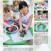 Two children sit outside, grinning as they paint corn cobs at a table filled with art supplies, inspired by activities from "Kinder Gardens" by Schiffer Publishing, Ltd. Painted corn and leaves complete this fun gardening scene.