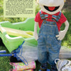 A smiling puppet in overalls and a red shirt stands with gardening tools, boots, and a container—Kinder Gardens by Schiffer Publishing, Ltd. inspires kids' gardening and outdoor play in a sunny garden.