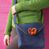 A person in a green sweater and brown pants holds "Felting" by Schiffer Publishing, Ltd.—a blue felted shoulder bag with an orange flower flap—standing before a pink wall.