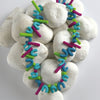 A Schiffer Publishing, Ltd. Costume Jewelry necklace with blue, green, and purple beads is displayed on smooth white stones, set against a white background with a red band at the top.